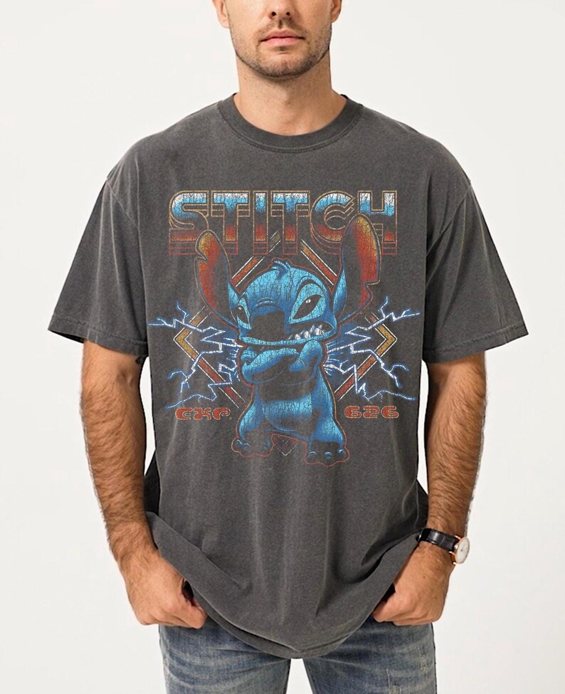 Peut inclure: T-shirt gris fonc&eacute; avec un graphisme du personnage Stitch de Lilo & Stitch. Le motif comprend le mot "Stitch" dans une police r&eacute;tro, Stitch les bras crois&eacute;s et des &eacute;clairs. Le t-shirt est un col rond.