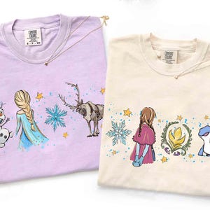 K&ouml;nnte beinhalten: Zwei T-Shirts mit Rundhalsausschnitt, eines in Helllila und eines in Creme, mit aufgedruckten Grafiken von Zeichentrickfiguren. Das lila Shirt zeigt einen Schneemann, eine blonde Figur und ein Rentier. Das cremefarbene Shirt zeigt eine Figur mit Z&ouml;pfen, ein florales Emblem und eine blaue Eidechse, beide akzentuiert mit blauen Schneeflocken und gelben Sternen.