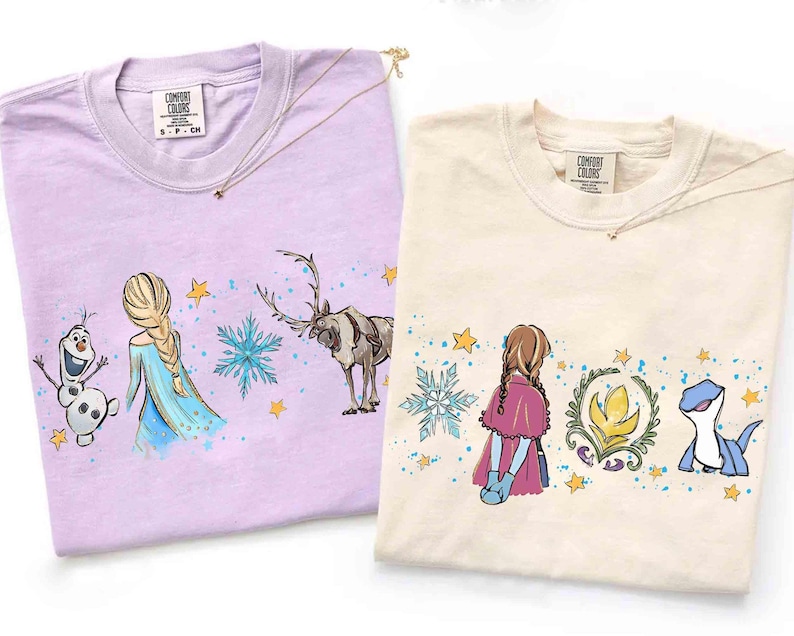 K&ouml;nnte beinhalten: Zwei T-Shirts mit Rundhalsausschnitt, eines in Helllila und eines in Creme, mit aufgedruckten Grafiken von Zeichentrickfiguren. Das lila Shirt zeigt einen Schneemann, eine blonde Figur und ein Rentier. Das cremefarbene Shirt zeigt eine Figur mit Z&ouml;pfen, ein florales Emblem und eine blaue Eidechse, beide akzentuiert mit blauen Schneeflocken und gelben Sternen.