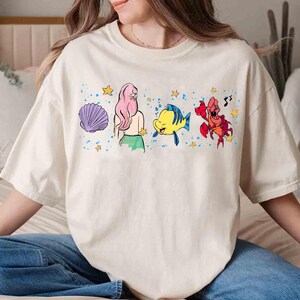 May include: A cream-colored short-sleeve t-shirt featuring a graphic print of a purple seashell, a mermaid with long pink hair, a yellow and blue fish, and a red crab, all surrounded by small yellow stars and blue paint splatter accents.