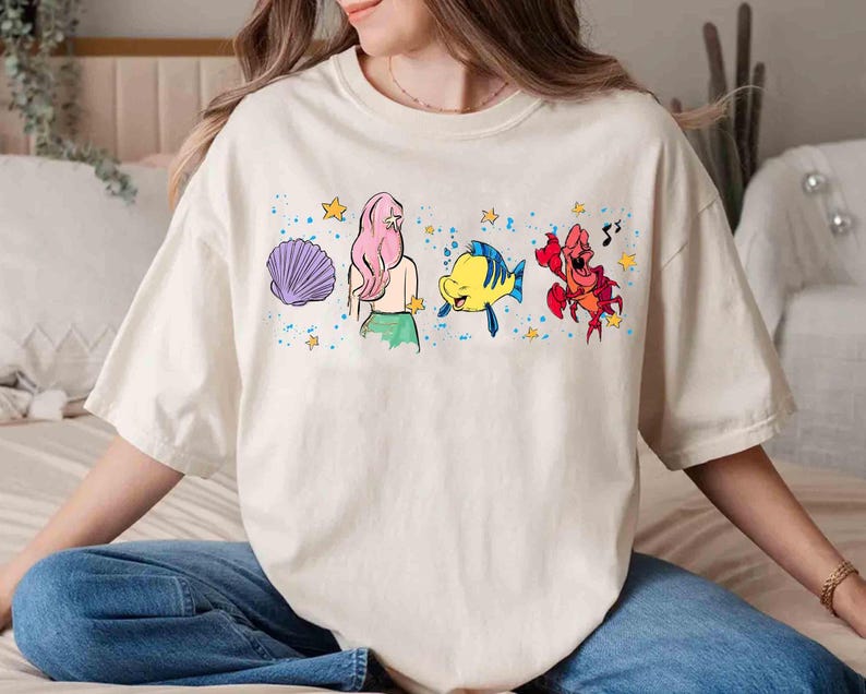 May include: A cream-colored short-sleeve t-shirt featuring a graphic print of a purple seashell, a mermaid with long pink hair, a yellow and blue fish, and a red crab, all surrounded by small yellow stars and blue paint splatter accents.
