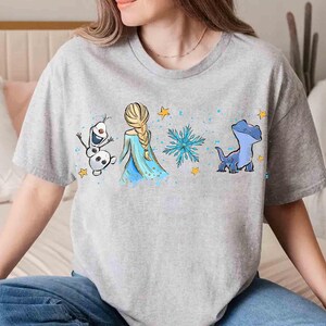 K&ouml;nnte beinhalten: Ein graues, meliertes Kurzarm-T-Shirt mit einem Grafikdruck, der einen wei&szlig;en Schneemann, eine blonde Figur mit langem Zopf in einem blauen Kleid, eine blaue Schneeflocke und ein kleines blaues Wesen zeigt, akzentuiert durch gelbe Sterne.