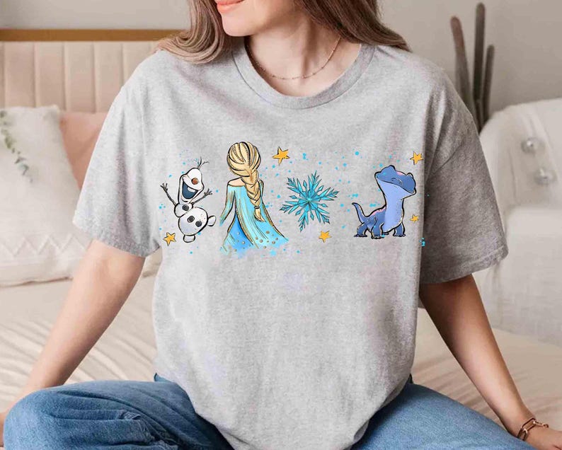 K&ouml;nnte beinhalten: Ein graues, meliertes Kurzarm-T-Shirt mit einem Grafikdruck, der einen wei&szlig;en Schneemann, eine blonde Figur mit langem Zopf in einem blauen Kleid, eine blaue Schneeflocke und ein kleines blaues Wesen zeigt, akzentuiert durch gelbe Sterne.