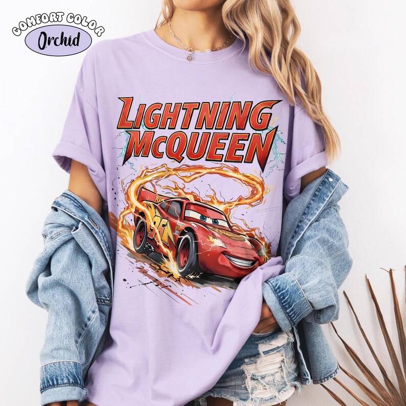 Op de afbeelding: Lichtpaars T-shirt met de tekst "LIGHTNING McQUEEN" in rood en geel. Het ontwerp toont een afbeelding van de rode raceauto Lightning McQueen met vlammen en bliksem. Het shirt is gelabeld "COMFORT COLOR Orchid".