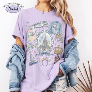 Puede incluir: Una camiseta lila Comfort Colors con un dise&ntilde;o de fantas&iacute;a que presenta un castillo, un tren y otros gr&aacute;ficos tem&aacute;ticos. La camiseta incluye el texto "Most Magical Place" y varias ilustraciones, incluyendo una corona y orejas de rat&oacute;n.