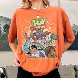 May include: Orange short-sleeved t-shirt featuring a graphic of characters from the animated film "Toy Story." The design includes the film's title and various characters such as Buzz Lightyear, Woody, and Rex. The shirt also has the names of the characters at the bottom.