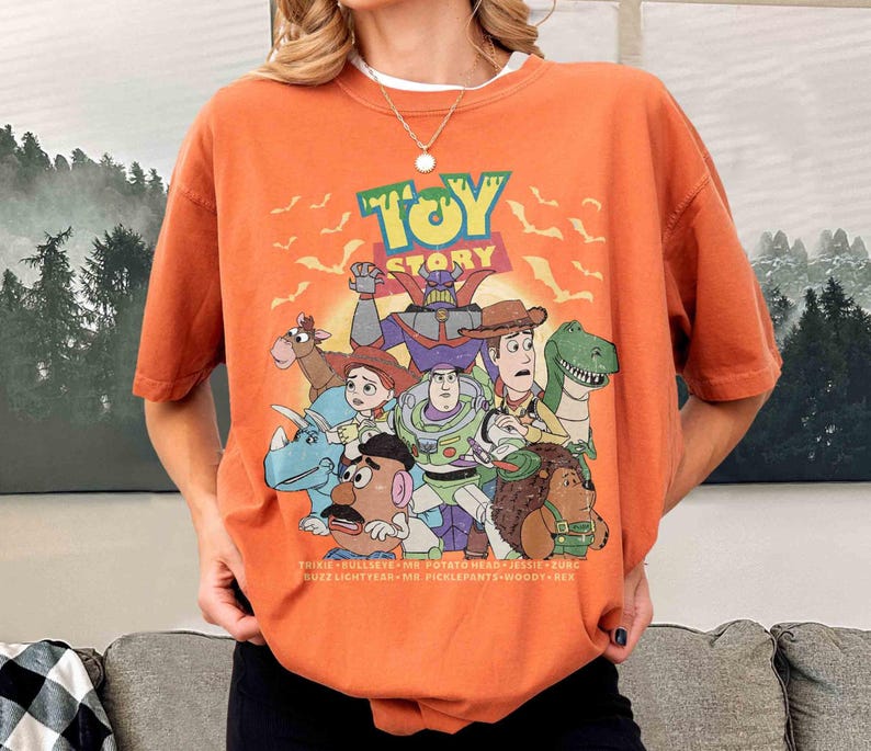 May include: Orange short-sleeved t-shirt featuring a graphic of characters from the animated film "Toy Story." The design includes the film's title and various characters such as Buzz Lightyear, Woody, and Rex. The shirt also has the names of the characters at the bottom.