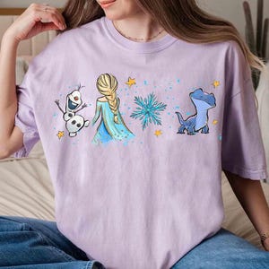 K&ouml;nnte beinhalten: Ein hellviolettes Kurzarm-T-Shirt mit einem Grafikdruck, der einen Schneemann, eine blonde Figur mit Zopf in einem blauen Kleid, eine blaue Schneeflocke und eine kleine blaue Eidechse zeigt, akzentuiert durch gelbe Sterne und blaue Farbspritzer.