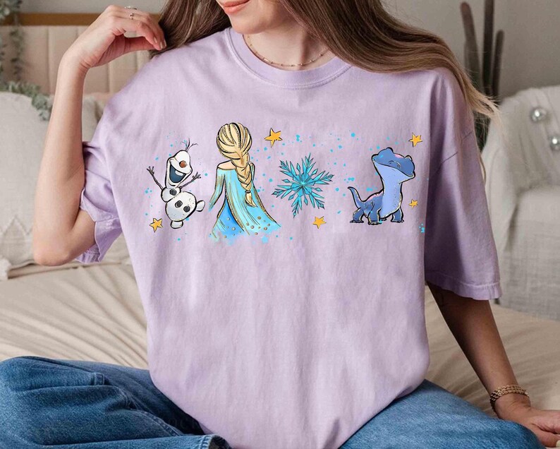 K&ouml;nnte beinhalten: Ein hellviolettes Kurzarm-T-Shirt mit einem Grafikdruck, der einen Schneemann, eine blonde Figur mit Zopf in einem blauen Kleid, eine blaue Schneeflocke und eine kleine blaue Eidechse zeigt, akzentuiert durch gelbe Sterne und blaue Farbspritzer.