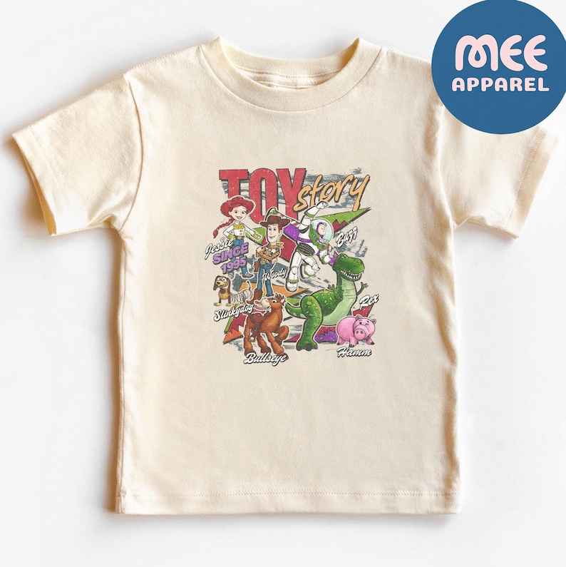 Pu&ograve; includere: Una t-shirt a maniche corte color crema con una stampa grafica dei personaggi di Toy Story, tra cui Woody, Buzz Lightyear, Jessie, Rex, Hamm, Bullseye e Slinky Dog. Il testo riporta Toy Story, Since 1995, e identifica ogni personaggio per nome.