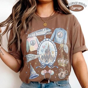 Puede incluir: Camiseta marr&oacute;n con una colorida ilustraci&oacute;n de un castillo, su&eacute;teres, llaves y otros iconos de parques tem&aacute;ticos. La camiseta tiene las palabras "Most Magical Place" y "Comfort Color" impresas.
