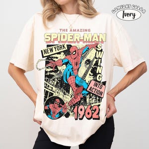 Puede incluir: Camiseta color marfil con un gr&aacute;fico vintage de Spider-Man. El dise&ntilde;o incluye las palabras "THE AMAZING SPIDER-MAN", "NEW YORK" y "1962". Spider-Man se representa con su traje cl&aacute;sico rojo y azul, balance&aacute;ndose. La camiseta est&aacute; etiquetada como "COMFORT COLOR".