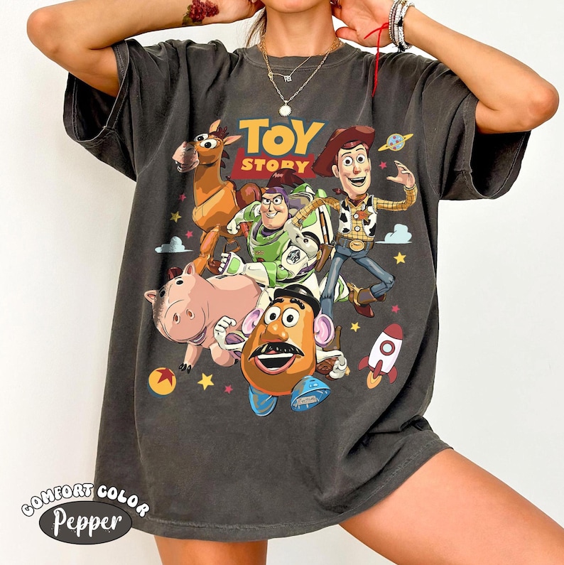 May include: Dark gray t-shirt featuring a graphic of characters from the animated film "Toy Story." The design includes Woody, Buzz Lightyear, Mr. Potato Head, Hamm, and Bullseye. The text "TOY STORY" is displayed above the characters. The shirt also has the text "Comfort Color Pepper".