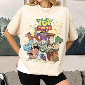 May include: Cream-colored t-shirt featuring the Toy Story logo and characters. The graphic includes Buzz Lightyear, Woody, and other characters from the animated film. The shirt has a relaxed fit and short sleeves.