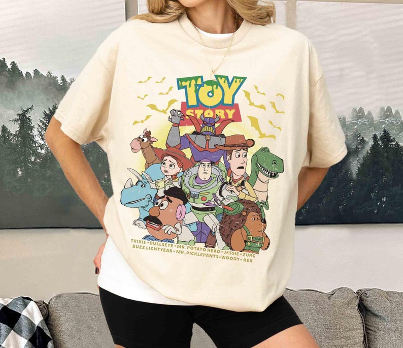 May include: Cream-colored t-shirt featuring the Toy Story logo and characters. The graphic includes Buzz Lightyear, Woody, and other characters from the animated film. The shirt has a relaxed fit and short sleeves.