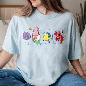May include: A light blue t-shirt featuring a graphic print of a mermaid with pink hair, a yellow fish, and a red crab, surrounded by purple seashells, yellow stars, and blue paint splatter accents across the center of the chest.