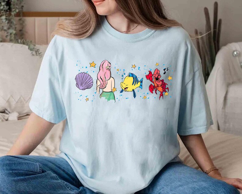 May include: A light blue t-shirt featuring a graphic print of a mermaid with pink hair, a yellow fish, and a red crab, surrounded by purple seashells, yellow stars, and blue paint splatter accents across the center of the chest.