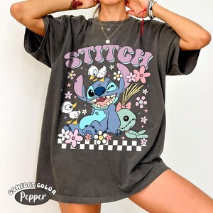 May include: Dark grey t-shirt with the word "STITCH" in a retro font above a cartoon image of Stitch, flowers, and ducks. The shirt has a comfort colour and pepper label.