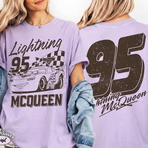 May include: A light purple t-shirt featuring a graphic of Lightning McQueen from the Cars movie. The front design includes the text "Lightning 95 McQueen" and an image of the race car. The back of the shirt displays a large "95" and "Lightning McQueen".