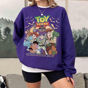 May include: A purple sweatshirt featuring the text "TOY STORY" above a graphic of cartoon characters. The characters include Woody, Buzz Lightyear, and Mr. Potato Head. The names of the characters are listed at the bottom of the graphic.
