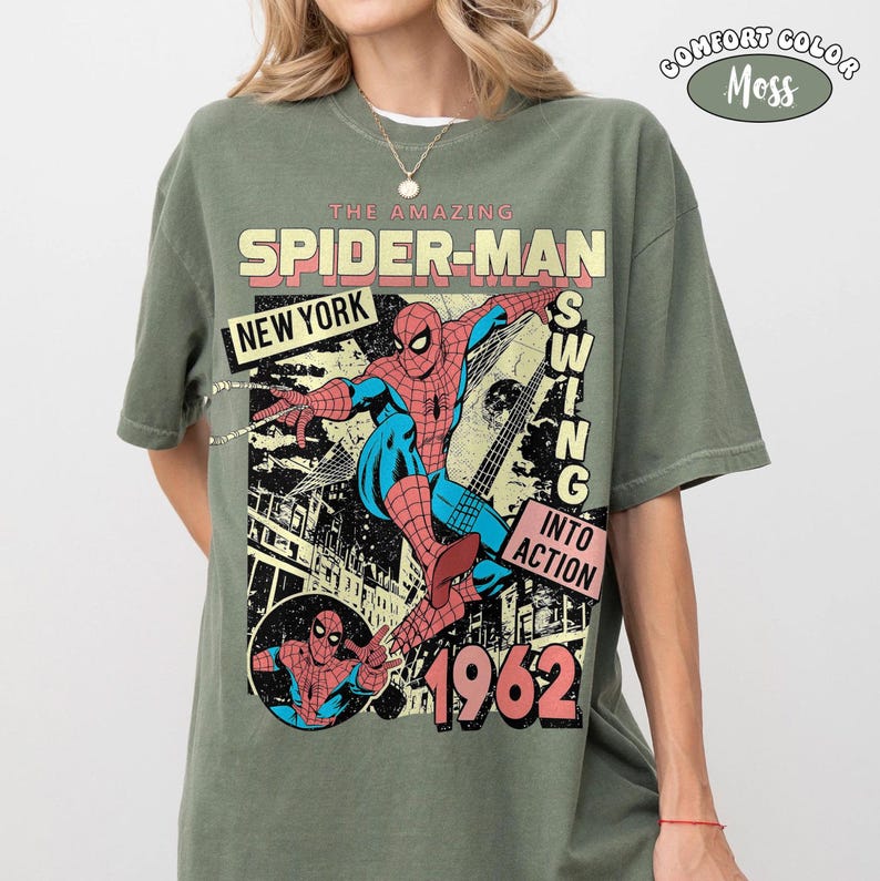 Puede incluir: Una camiseta verde musgo con un gr&aacute;fico de Spider-Man en acci&oacute;n. El dise&ntilde;o incluye el texto "THE AMAZING SPIDER-MAN NEW YORK SWING INTO ACTION 1962". La camiseta est&aacute; etiquetada como "COMFORT COLOR Moss".