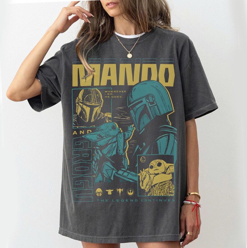 Pode incluir: T-shirt cinzenta escura com um design gr&aacute;fico com o texto "MANDO AND GROGU". O design inclui imagens de um personagem com capacete e uma pequena criatura verde. O texto "THE LEGEND CONTINUES" tamb&eacute;m &eacute; vis&iacute;vel.
