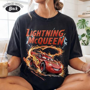 Op de afbeelding: Zwart Comfort Colors T-shirt met Lightning McQueen uit de film Cars. De afbeelding toont de rode raceauto met de tekst "Lightning McQueen" in rood en geel. Het shirt heeft een vintage stijl.