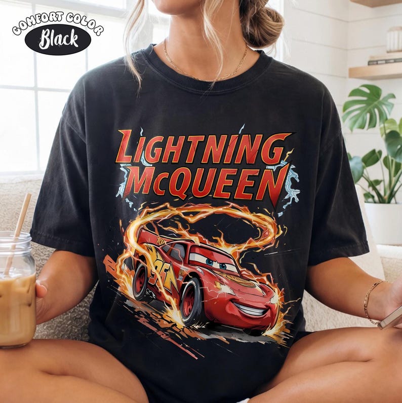 Op de afbeelding: Zwart Comfort Colors T-shirt met Lightning McQueen uit de film Cars. De afbeelding toont de rode raceauto met de tekst "Lightning McQueen" in rood en geel. Het shirt heeft een vintage stijl.