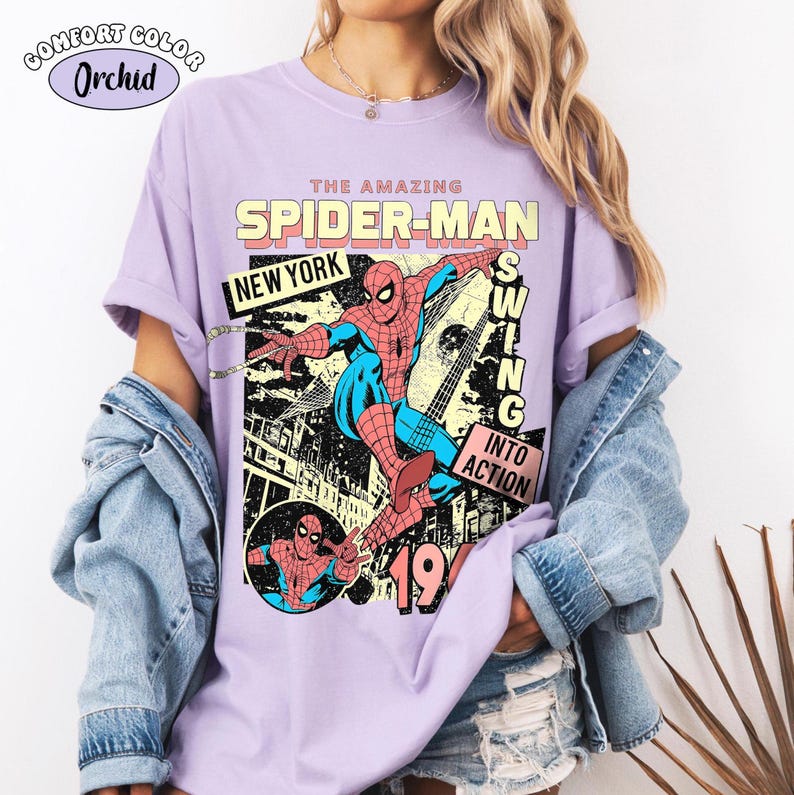 Puede incluir: Una camiseta de color lila claro con un gr&aacute;fico de c&oacute;mic de Spider-Man. El dise&ntilde;o incluye el texto "THE AMAZING SPIDER-MAN", "NEW YORK" y "SWING INTO ACTION". La camiseta tambi&eacute;n tiene el texto "COMFORT COLOR Orchid".