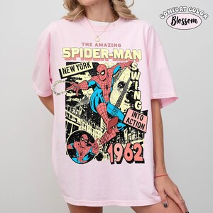 Puede incluir: Camiseta rosa claro Comfort Colors con un dise&ntilde;o de c&oacute;mic vintage de Spider-Man. El dise&ntilde;o incluye el texto "THE AMAZING SPIDER-MAN", "NEW YORK", "SWING INTO ACTION" y el a&ntilde;o "1962". La camiseta tambi&eacute;n tiene el logo "Comfort Color Blossom".