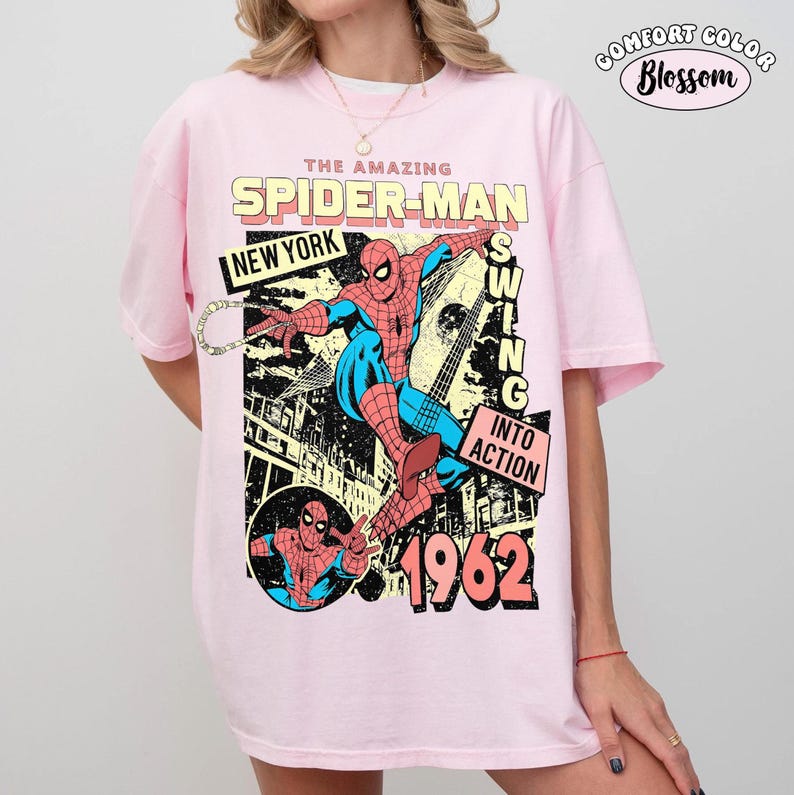 Puede incluir: Camiseta rosa claro Comfort Colors con un dise&ntilde;o de c&oacute;mic vintage de Spider-Man. El dise&ntilde;o incluye el texto "THE AMAZING SPIDER-MAN", "NEW YORK", "SWING INTO ACTION" y el a&ntilde;o "1962". La camiseta tambi&eacute;n tiene el logo "Comfort Color Blossom".