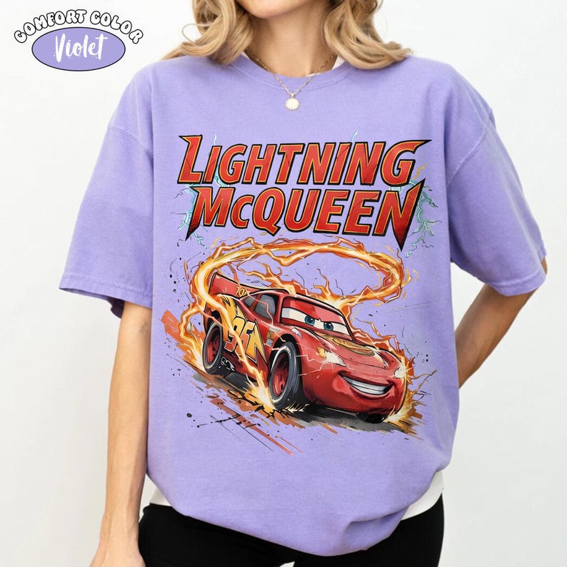 Op de afbeelding: Een violet Comfort Colors T-shirt met een afbeelding van Lightning McQueen uit de film Cars. De rode raceauto is omringd door vlammen en de tekst "Lightning McQueen" in rood en geel.