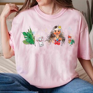 May include: A light pink short-sleeve t-shirt featuring a watercolor-style graphic print. The design includes a green tropical leaf, a small white pig, a female character with long brown hair and a yellow flower, and a colorful rooster character.