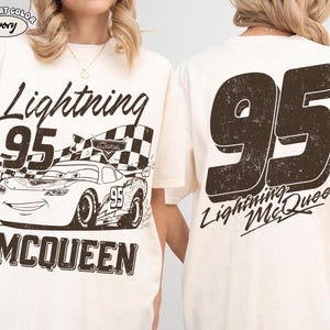 May include: Ivory-colored t-shirt featuring a distressed graphic of Lightning McQueen from the movie Cars. The front displays the text "Lightning 95 McQueen" with an image of the car and a checkered flag. The back has a large "95" and "Lightning McQueen".
