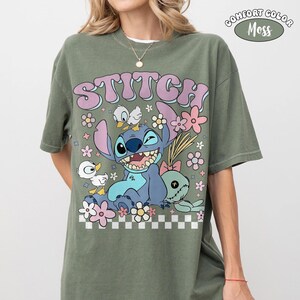 May include: Olive green t-shirt with a Stitch graphic from Lilo & Stitch, flowers, and ducks. The word "STITCH" is in bubble letters. The shirt is labelled "Comfort Color Moss".