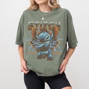 Peut inclure: T-shirt vert olive avec un motif de Stitch de Lilo & Stitch. Le design comprend le mot "STITCH", l'image du personnage et les chiffres "626". Le t-shirt a des manches courtes et une coupe d&eacute;contract&eacute;e.