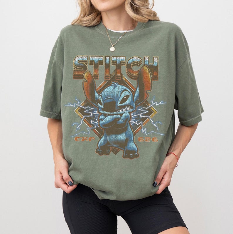 Peut inclure: T-shirt vert olive avec un motif de Stitch de Lilo & Stitch. Le design comprend le mot "STITCH", l'image du personnage et les chiffres "626". Le t-shirt a des manches courtes et une coupe d&eacute;contract&eacute;e.
