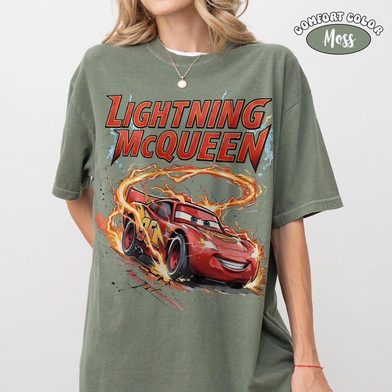 Op de afbeelding: Een mosgroen t-shirt met de tekst "LIGHTNING McQUEEN" in rood en geel, en een afbeelding van de rode raceauto. Het shirt is van het merk Comfort Colors.