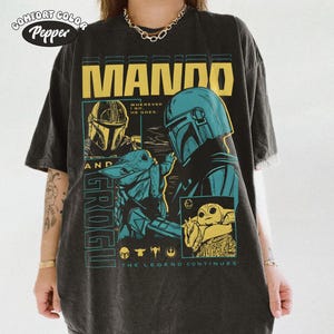 Pode incluir: Camiseta cinza escuro com um design gr&aacute;fico com o texto "MANDO" em letras amarelas. O design inclui ilustra&ccedil;&otilde;es de The Mandalorian e Grogu, com o texto "THE LEGEND CONTINUES". A camiseta tem um visual vintage e desgastado.