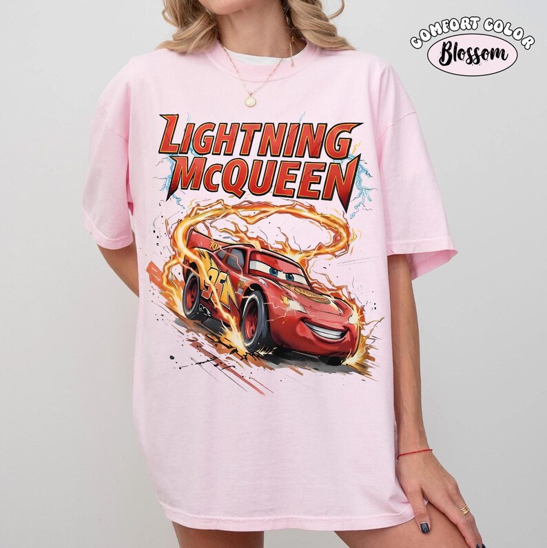 Op de afbeelding: Lichtroze T-shirt met een afbeelding van Lightning McQueen uit de film Cars. Het ontwerp bevat de tekst "LIGHTNING McQUEEN" in rood en geel, met het personage in beweging, omringd door vlammen.