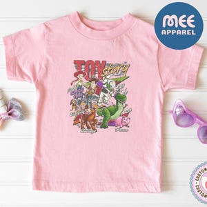 Pu&ograve; includere: Una t-shirt a maniche corte rosa chiaro con una stampa grafica colorata dei personaggi di Toy Story, tra cui Woody, Buzz Lightyear, Jessie, Rex, Hamm, Bullseye e Slinky Dog, con il testo Toy Story, Jessie Since 1995 e i nomi dei personaggi.