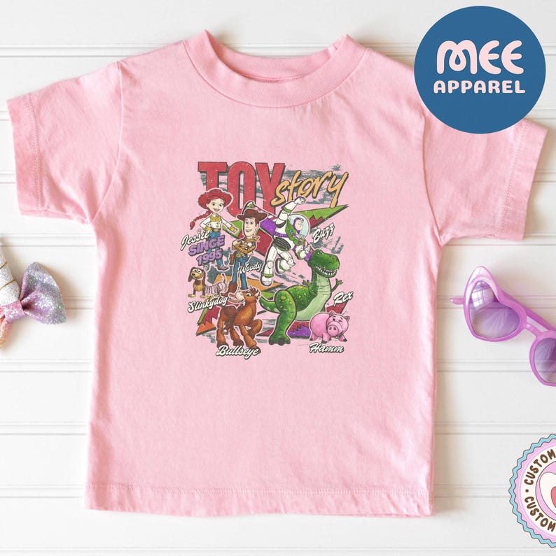 Pu&ograve; includere: Una t-shirt a maniche corte rosa chiaro con una stampa grafica colorata dei personaggi di Toy Story, tra cui Woody, Buzz Lightyear, Jessie, Rex, Hamm, Bullseye e Slinky Dog, con il testo Toy Story, Jessie Since 1995 e i nomi dei personaggi.