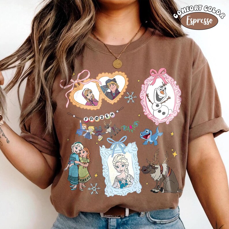 May include: Espresso-colored t-shirt featuring various characters and elements from the animated film "Frozen." The design includes framed portraits of characters, snowflakes, and the word "FROZEN" in a banner. The shirt has a Comfort Color label.