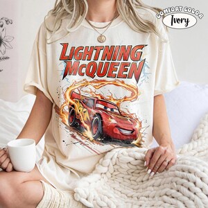 Op de afbeelding: Ivoor kleurig T-shirt met een afbeelding van Lightning McQueen uit de film Cars. Het ontwerp bevat de tekst "Lightning McQueen" in het rood en een rode raceauto met nummer 95. Het shirt is gelabeld "Comfort Color Ivory".
