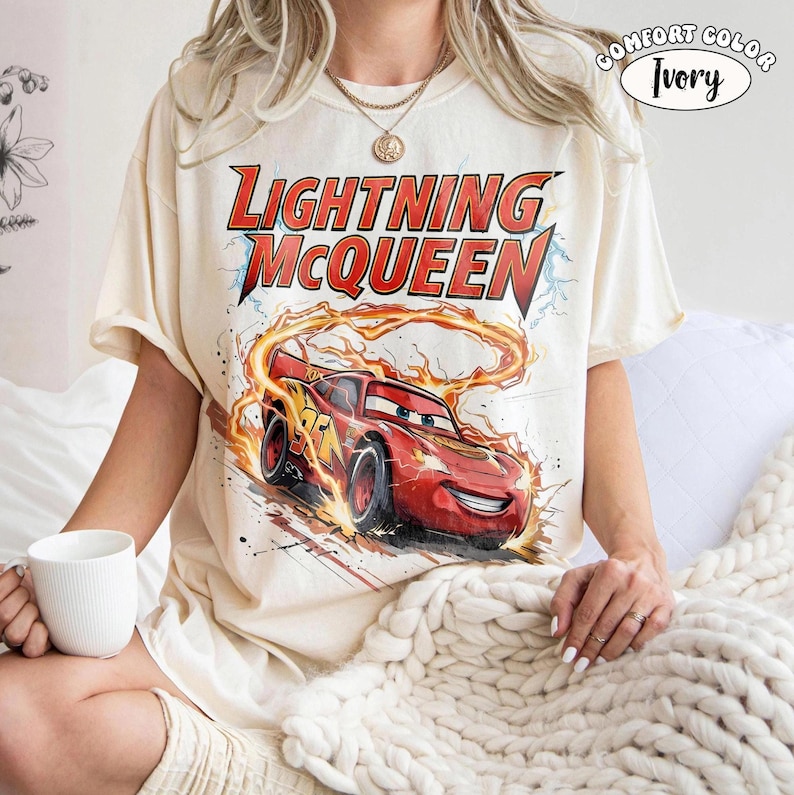 Op de afbeelding: Ivoor kleurig T-shirt met een afbeelding van Lightning McQueen uit de film Cars. Het ontwerp bevat de tekst "Lightning McQueen" in het rood en een rode raceauto met nummer 95. Het shirt is gelabeld "Comfort Color Ivory".