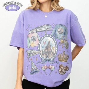 Puede incluir: Una camiseta violeta Comfort Colors con una ilustraci&oacute;n caprichosa de un castillo, un tren, llaves y otros iconos de parques tem&aacute;ticos. La camiseta incluye el texto "Most Magical Place" en una pancarta y una galleta de Mickey Mouse.