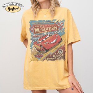 May include: Mustard-coloured t-shirt featuring a graphic of Lightning McQueen from the film Cars. The design includes text that reads "Lightning McQueen" and "Winner Takes All." The shirt is a Comfort Color brand.