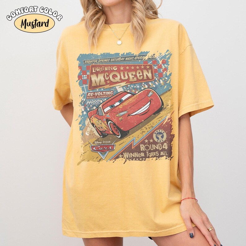 May include: Mustard-coloured t-shirt featuring a graphic of Lightning McQueen from the film Cars. The design includes text that reads "Lightning McQueen" and "Winner Takes All." The shirt is a Comfort Color brand.