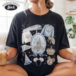 Puede incluir: Camiseta negra Comfort Colors con un gr&aacute;fico colorido de un castillo, su&eacute;teres, un tren y otros elementos tem&aacute;ticos. La camiseta incluye el texto "Most Magical Place" en una bandera y una galleta de Mickey Mouse.
