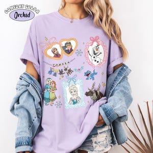 May include: A light purple t-shirt with multiple cartoon images from the movie "Frozen." The shirt features images of characters like Elsa, Anna, Olaf, and Kristoff, framed in decorative borders. The word "FROZEN" is printed in a playful font.